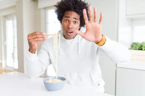 African American man eating asian noodles using chopsticks at home with open hand doing stop sign with serious and confident expression, defense gesture