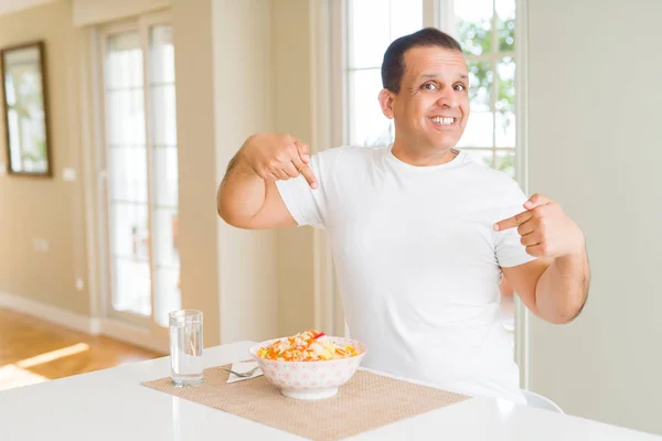 Middle age man eating rice at home looking confident with smile on face, pointing oneself with fingers proud and happy.