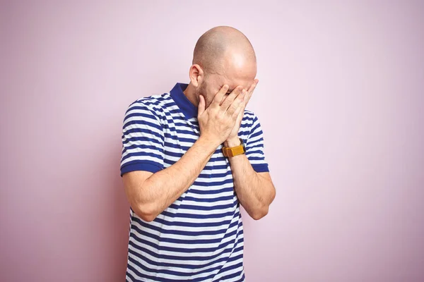 Young bald man with beard wearing casual striped blue t-shirt over pink isolated background with sad expression covering face with hands while crying. Depression concept.