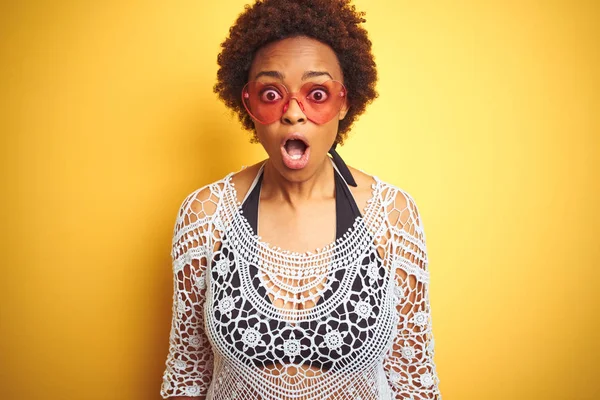 Young african american woman with afro hair wearing bikini and heart shaped sunglasses afraid and shocked with surprise expression, fear and excited face.