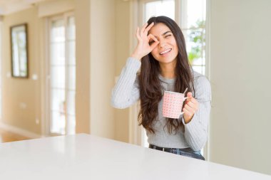 Mutlu yüzü ile bir fincan kahve içen genç kadın parmaklarından bakarak el iyle ok işareti yapıyor gülümseyerek