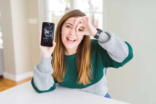 Beautiful young woman holding broken smartphone with happy face smiling doing ok sign with hand on eye looking through fingers