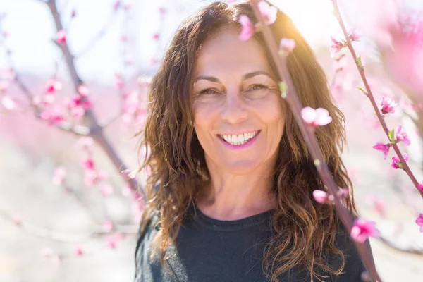 Beautiful middle age woman in the middle of pink peach flowers and trees smiling cheerful, colorful sunny day of spring