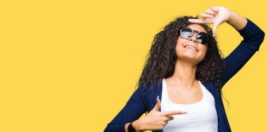 Genç güzel kız moda elle yapma çerçeve ve parmakları ile mutlu yüz gülümseyen güneş gözlüğü takıyor kıvırcık saçlı. Yaratıcılık ve Fotoğrafçılık kavramı.