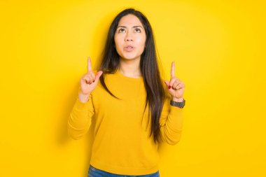 Sarı izole arka plan üzerinde güzel esmer kadın şaşırttı ve yukarı bakarak ve parmakları ile işaret ve kolları kaldırdı şaşırttı.