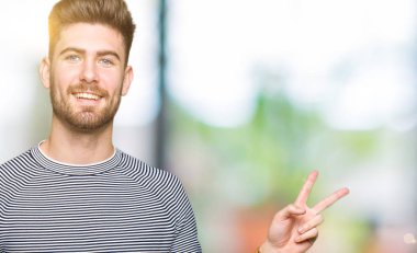 Çizgili kazak giyen genç yakışıklı adam mutlu yüz le gülümseyerek zafer işareti yaparak kameraya göz kırpıyor. İki numara.