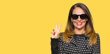 Güzel orta yaş kadın mutlu yüz zafer işareti yaparken kameraya göz kırpıyor ile gülümseyen güneş gözlüğü takıyor. İki numaralı.