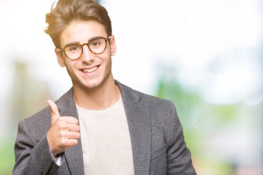 İzole arka planda gözlük takan genç iş adamı el ile jest kadar mutlu başparmak yapıyor. Başarı gösteren kameraya bakarak ifadeyi onaylama.