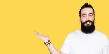 Uzun saç ve sakal neşeli sunulması ve kameraya bakarak avucunun ile işaret gülümseyen casual beyaz t-shirt giyen genç hipster adamla.