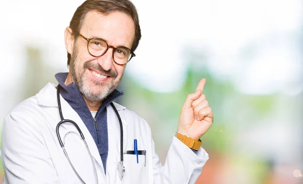 カメラを見て横に手と指で指差し 顔に大きな笑顔を持つ医療コートを着たハンサムな中年の医者の男 — ストック写真