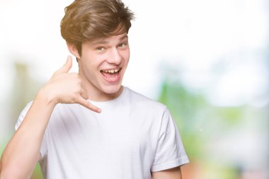 Genç yakışıklı adam giyiyor rahat beyaz tişört yapıyor gülümseyen izole arka plan üzerinde telefon hareketi ile el ve parmak telefonda konuşuyor gibi. Kavramları iletişim.