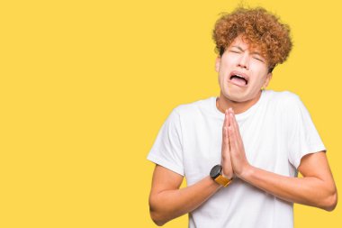 Afro saç ile genç yakışıklı adam rahat beyaz t-shirt yalvarıyor ve ellerle birlikte dua yüz çok duygusal ve endişeli karşısında umut ifade giyiyor. Affedilmesini istiyorum. Din konsepti.