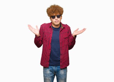Yakışıklı genç afro saçlı güneş gözlüğü clueless ve şaşkın bir ifadeyle kollar ve eller kaldırdı giyiyor. Şüphe kavramı.