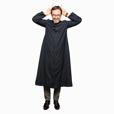 Katolik giyen orta yaş rahip adam robe komik poz ve baş parmak ile deli tavşan kulakları neşeli gülümseyen