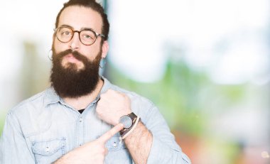 Uzun saç ve sakal acele işaretleme izlemek için zaman, sabırsızlık, üzgün ve kızgın son tarih gecikme için gözlük takan genç hipster adamla