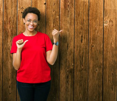 Güzel genç Afro-Amerikan kadın üzerinde gözlük takan izole arka plan emin gülümseyen arkasından el ve kadar başparmak ile işaret