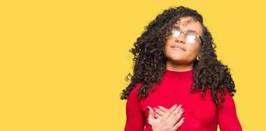 Kıvırcık saçlı gözlük ile gülümseyen genç güzel kadın kapalı gözler ve yüzünde minnettar jest ile göğüs üzerinde eller. Sağlık kavramı.