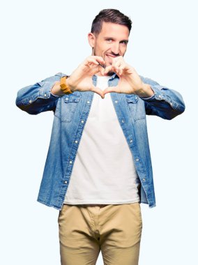 Mavi gözlü ve sakallı, kot ceket giyen yakışıklı adam sevgiyle gülümseyerek kalp sembolü ve şeklini elleriyle gösteriyor. Romantik kavram.