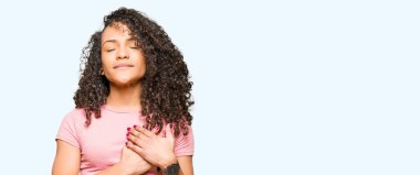 Kıvırcık saçlı pembe t-shirt ile gülümseyen giyen genç güzel kadın kapalı gözler ve yüzünde minnettar jest ile göğüs üzerinde eller. Sağlık kavramı.