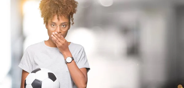 Young african american woman holding soccer football ball over isolated background cover mouth with hand shocked with shame for mistake, expression of fear, scared in silence, secret concept