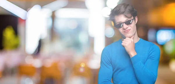 Young man wearing funny thug life glasses over isolated background with hand on chin thinking about question, pensive expression. Smiling with thoughtful face. Doubt concept.