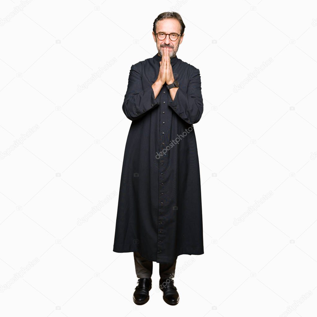 Middle age priest man wearing catholic robe praying with hands together asking for forgiveness smiling confident.