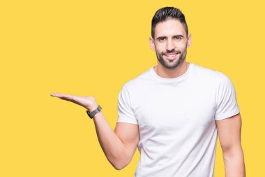 Neşeli sunulması ve kameraya bakarak avucunun ile işaret gülümseyen izole arka plan üzerinde rahat beyaz t-shirt giyen genç adam.