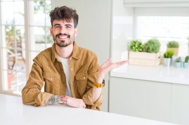 Beyaz masanın üzerinde oturan rahat ceket giyen genç adam neşeli sunum gülümseyerek ve kameraya bakarak avuç içi ile işaret.