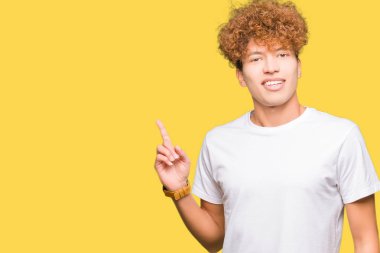 Kameraya bakarak yan el ve parmak ile işaret eden genç yakışıklı adam, yüzünde kocaman bir gülümseme ile rahat beyaz t-shirt giyen afro saçlı.