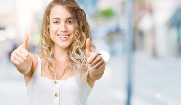 Young beautiful blonde woman over isolated background approving doing positive gesture with hand, thumbs up smiling and happy for success. Looking at the camera, winner gesture.