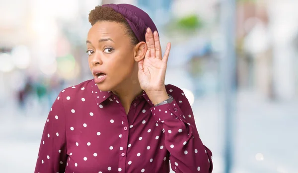 Beautiful young african american woman wearing head scarf over isolated background smiling with hand over ear listening an hearing to rumor or gossip. Deafness concept.