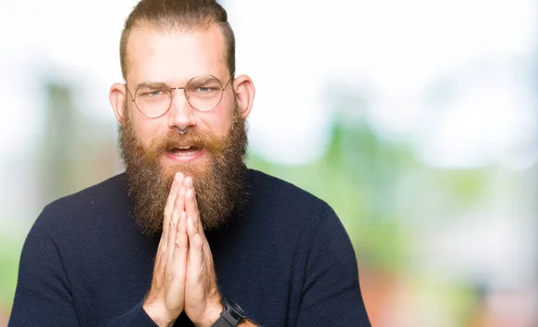 Young blond man wearing glasses and turtleneck sweater begging and praying with hands together with hope expression on face very emotional and worried. Asking for forgiveness. Religion concept.