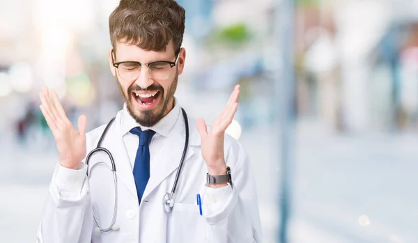 年轻的医生人穿医院外套在孤立的背景庆祝疯狂和疯狂为成功与胳膊举起和闭上眼睛尖叫兴奋 优胜者概念 — 图库照片
