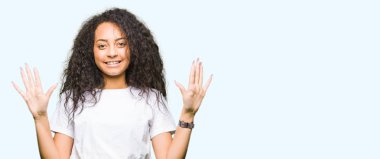 Rahat beyaz t-shirt gösteren ve parmak numarası on ile işaret giyen kıvırcık saçlı Genç güzel kız emin ve mutlu gülümseyen iken.
