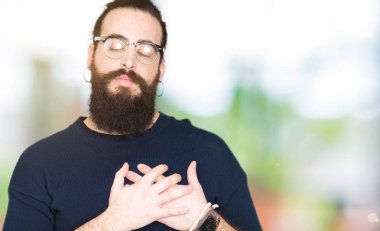 Uzun saç ve sakal ile gülümseyen gözlüklü genç hipster adamla kapalı gözler ve yüzünde minnettar jest ile göğüs üzerinde eller. Sağlık kavramı.