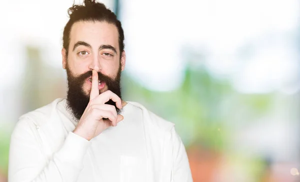 Young man with long hair and beard wearing sporty sweatshirt asking to be quiet with finger on lips. Silence and secret concept.