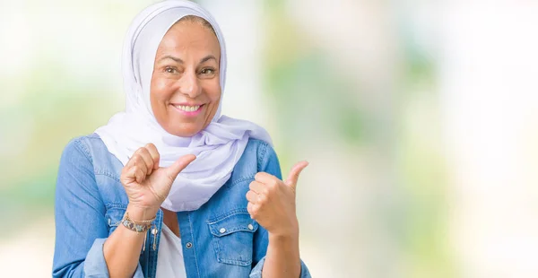 Middle age eastern arab woman wearing arabian hijab over isolated background Pointing to the back behind with hand and thumbs up, smiling confident
