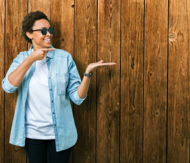 Güzel genç Afro-Amerikan kadın hayrete ve el ile sunulması ve parmak ile işaret ederken kameraya gülümseyen izole arka plan üzerinde güneş gözlüğü takıyor.