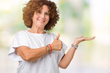 İzole arka plan gösteren palm el üzerinde beyaz t-shirt giyen ve yukarıya, mutlu ve neşeli gülümseyen baş parmakları Tamam jest yapıyor güzel orta ager üst düzey kadın