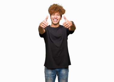 Afro saçlı siyah t-shirt onaylama giyen genç yakışıklı adam el ile pozitif jest yapıyor gülümseyen ve başarı için mutlu yaşasın. Kamera, kazanan jest arıyorsunuz.