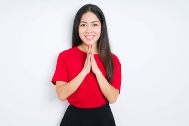 İzole arka plan üzerinde kırmızı t-shirt giyen güzel esmer kadın birlikte emin gülümseyerek af isteyen elleriyle dua.