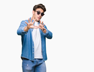 İzole arka plan üzerinde güneş gözlüğü takan genç yakışıklı adam korkuyor ve korku ifadesi ile korkuyor el ile jest durdurma, şok bağırıyor. Panik konsepti.