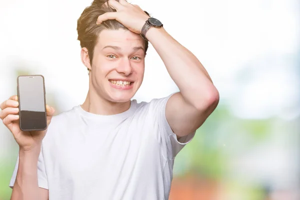 Young man showing smartphone screen over isolated background stressed with hand on head, shocked with shame and surprise face, angry and frustrated. Fear and upset for mistake.