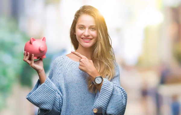 年轻美丽的金发碧眼的女人抱着小猪银行在孤立的背景非常高兴用手和手指指着 — 图库照片