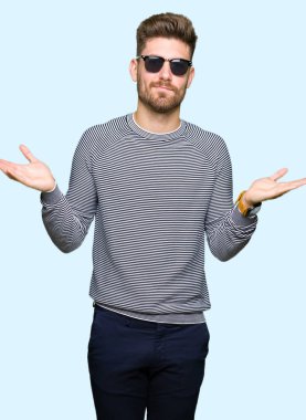 Güneş gözlüğü clueless ve şaşkın bir ifadeyle kollar ve eller kaldırdı giyen genç yakışıklı adam. Şüphe kavramı.