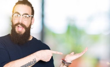Uzun saç ve sakal hayrete ve el ile sunulması ve parmak ile işaret ederken kameraya gülümseyen gözlüklü genç hipster adamla.