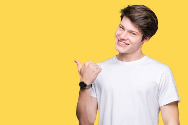 Seyir ve başparmak yanında yukarıyı mutlu yüz ile gülümseyen izole arka plan üzerinde rahat beyaz t-shirt giyen genç yakışıklı adam.