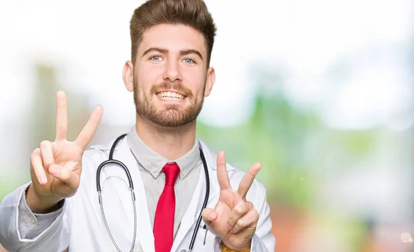 年轻英俊的医生穿着医疗外套微笑着看着镜头 显示手指做胜利的标志 — 图库照片