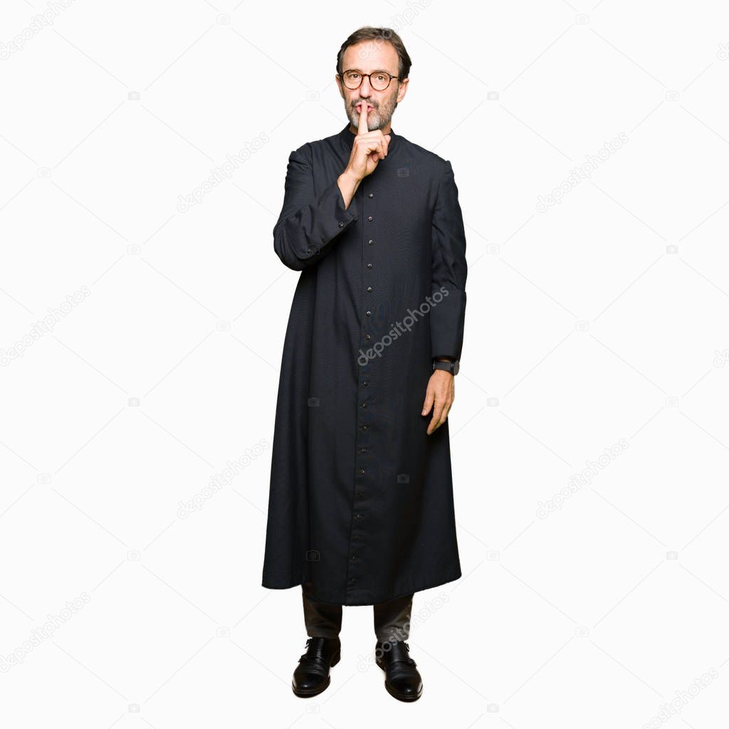 Middle age priest man wearing catholic robe asking to be quiet with finger on lips. Silence and secret concept.