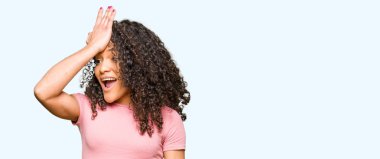 Young beautiful woman with curly hair wearing pink t-shirt surprised with hand on head for mistake, remember error. Forgot, bad memory concept. clipart